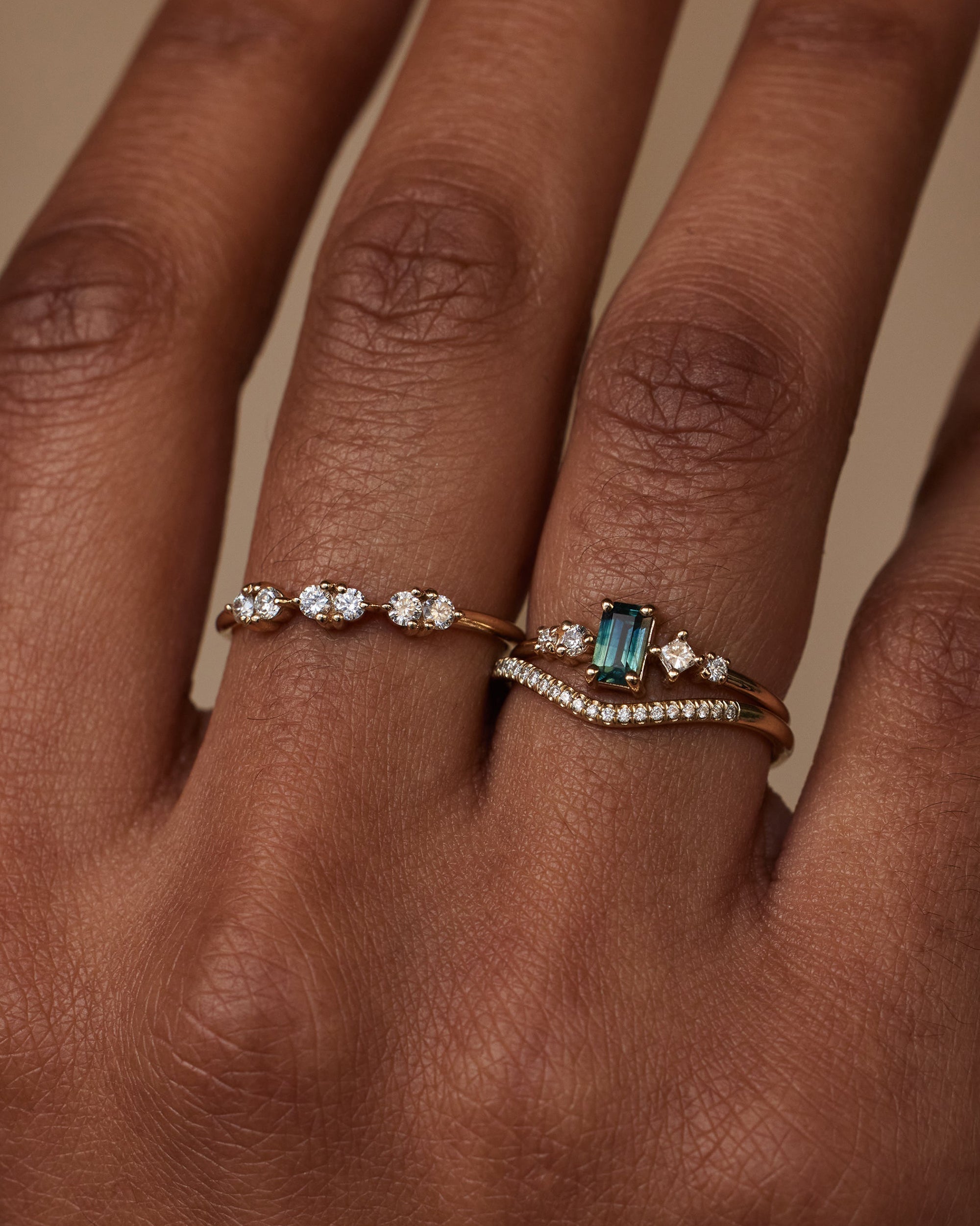 [lifestyle]Close-up of a hand wearing two diamond rings with a green gemstone on a neutral background
