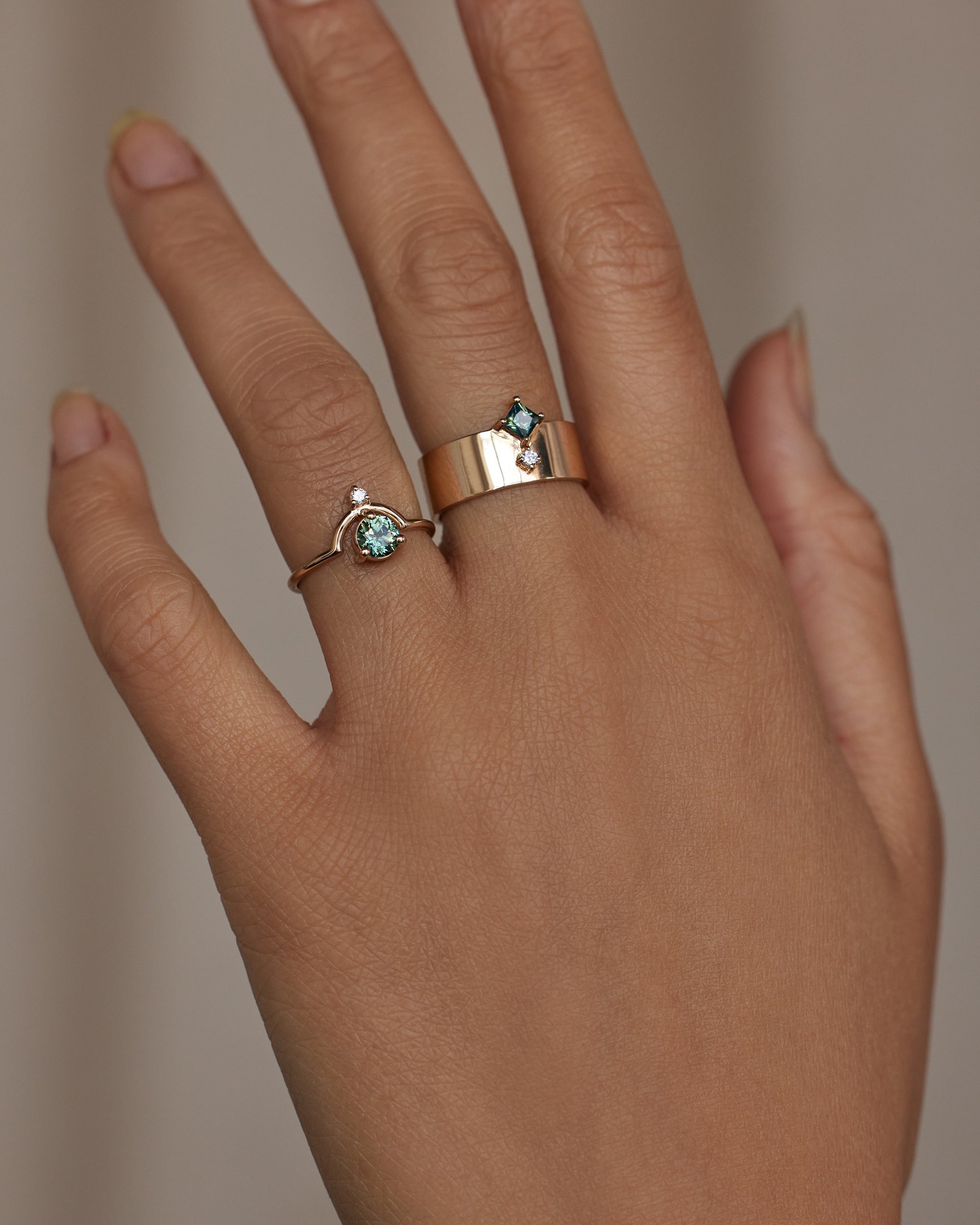 [lifestyle]Hand wearing two gold rings with green gemstones on a neutral background